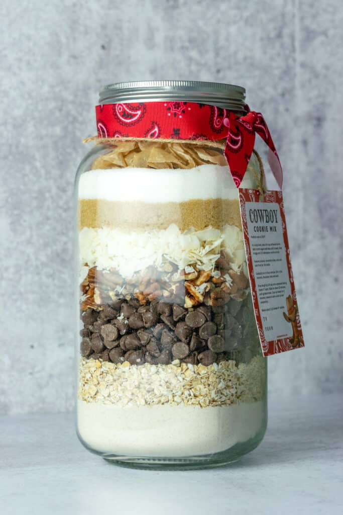 A neatly layered mason jar full of cowboy cookie mix with a gift tag hanging on it.