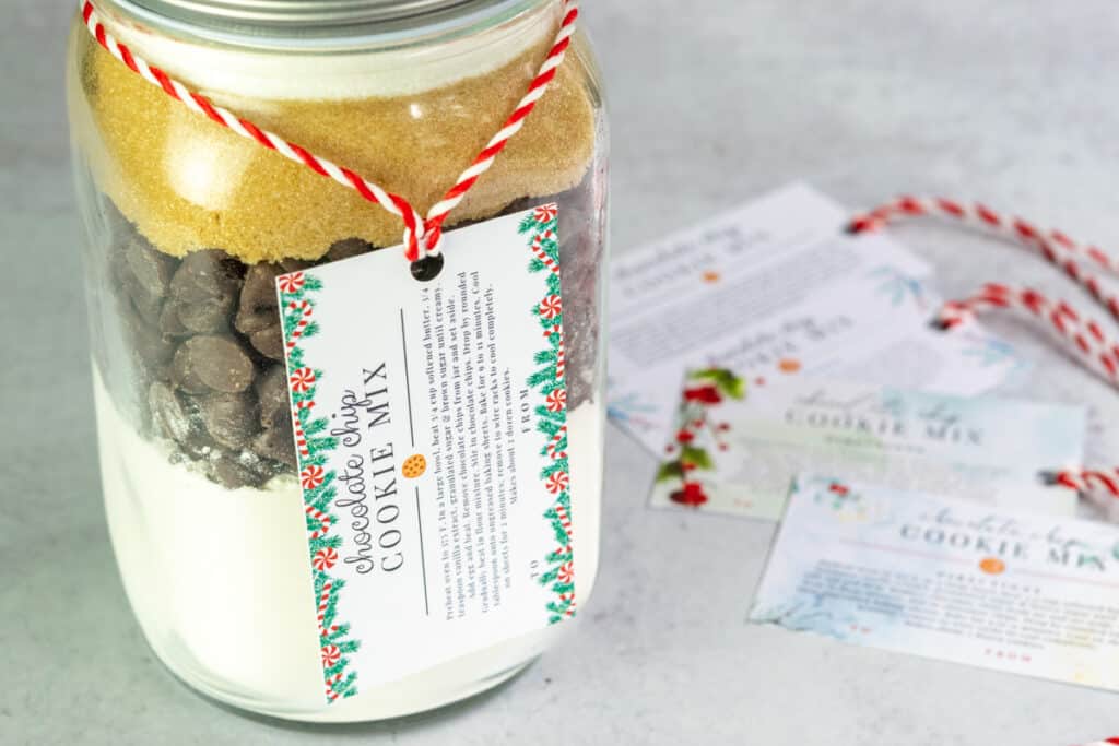 Chocolate chip cookie mix in a jar with a holiday gift tag hanging on it and more gift tags and red and white ribbon sitting next to the jar.