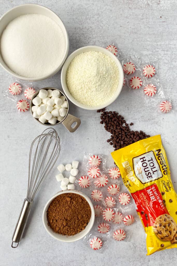 All the ingredients needed to put together a pint size jar of peppermint hot chocolate mix, including cocoa powder, powdered milk, chocolate chips spilling out of a bag, sugar, peppermint candies and marshmallows.