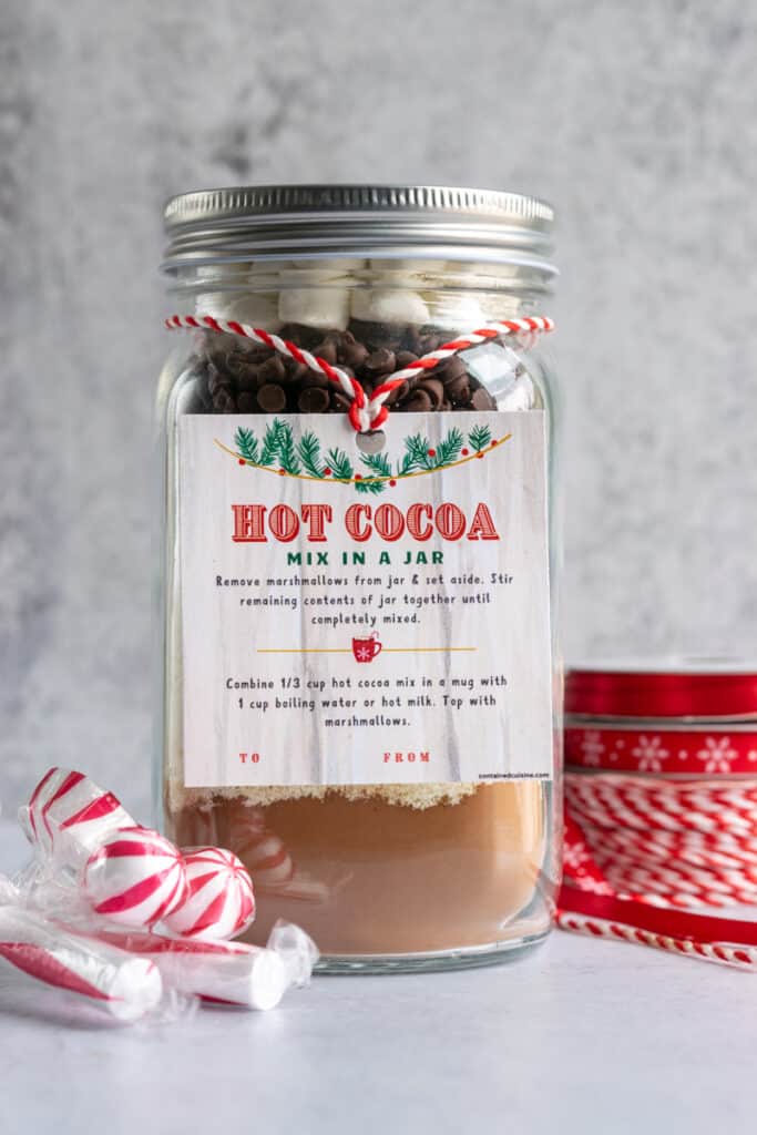Gift jar of hot cocoa mix with spools of snowflake and red and white striped ribbon next to it.