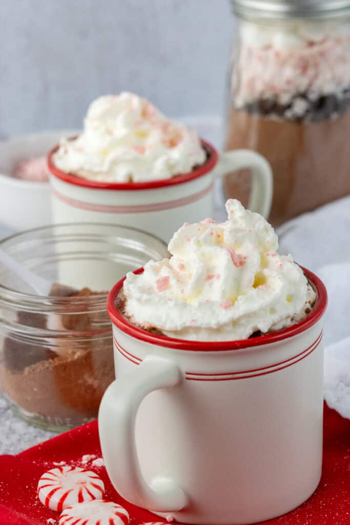 A jar of homemade peppermint hot cocoa with a scoop in it and two cups of hot chocolate topped with whipped cream and finely crushed peppermint.