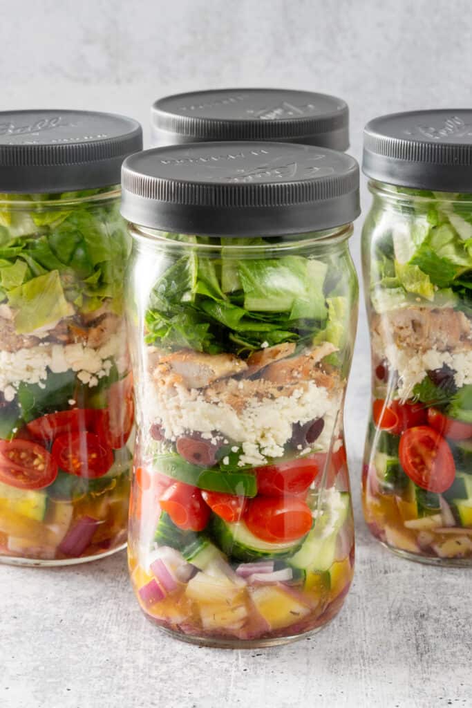 Four Greek salad mason jars layered with Greek dressing, veggies, olives, feta cheese and chicken.