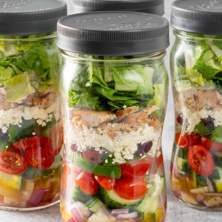 Four Greek salad mason jars layered with Greek dressing, veggies, olives, feta cheese and chicken.