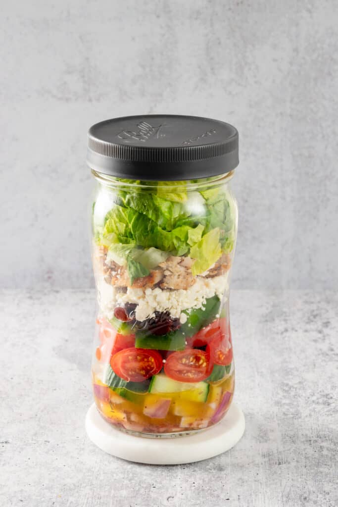 Greek salad layered in a tall quart size mason jar beginning with the dressing and onions and ending with lettuce.