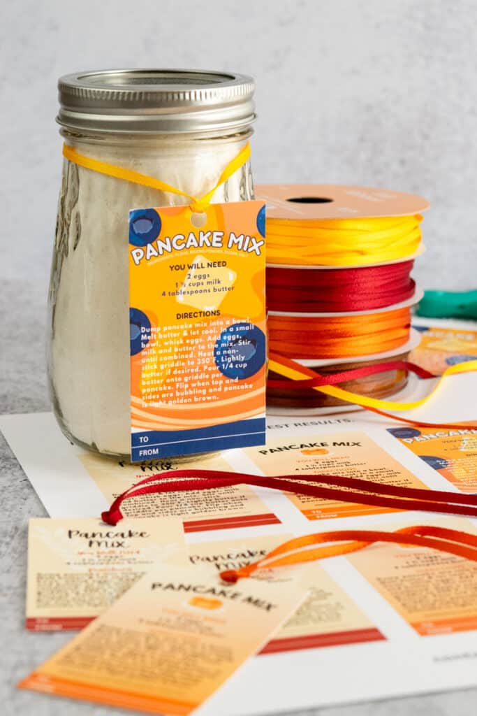 A pancake mix printable gift tag tied onto a jar which is sitting on top of printed gift tags and next to a spool of colorful ribbon.