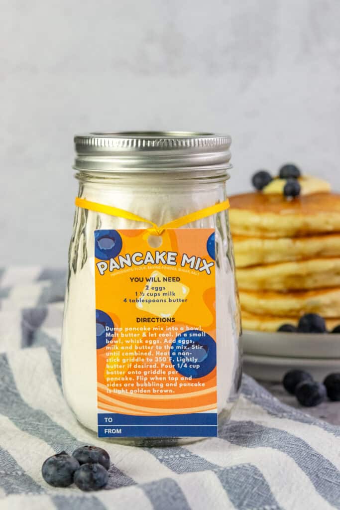 Pancake mix in a pint size mason jar with a gift tag sitting in front of a stack of fluffy pancakes that has butter, syrup and blueberries on top.