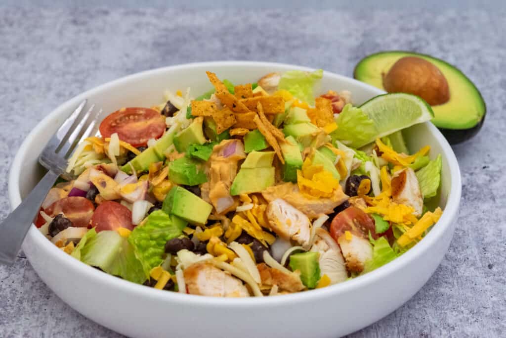 Large bowl of southwest salad with grilled chicken, onion, tomatoes, black beans, corn, cheese and diced avocado sitting in front of half of an avocado with pit in it.