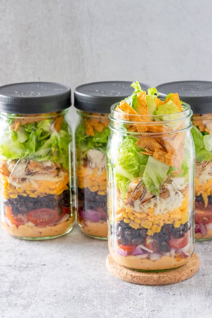 Four southwest salads in quart size salads where 3jars have leakproof lids screwed on and 1 jar is open with lettuce and tortilla strips sticking out.