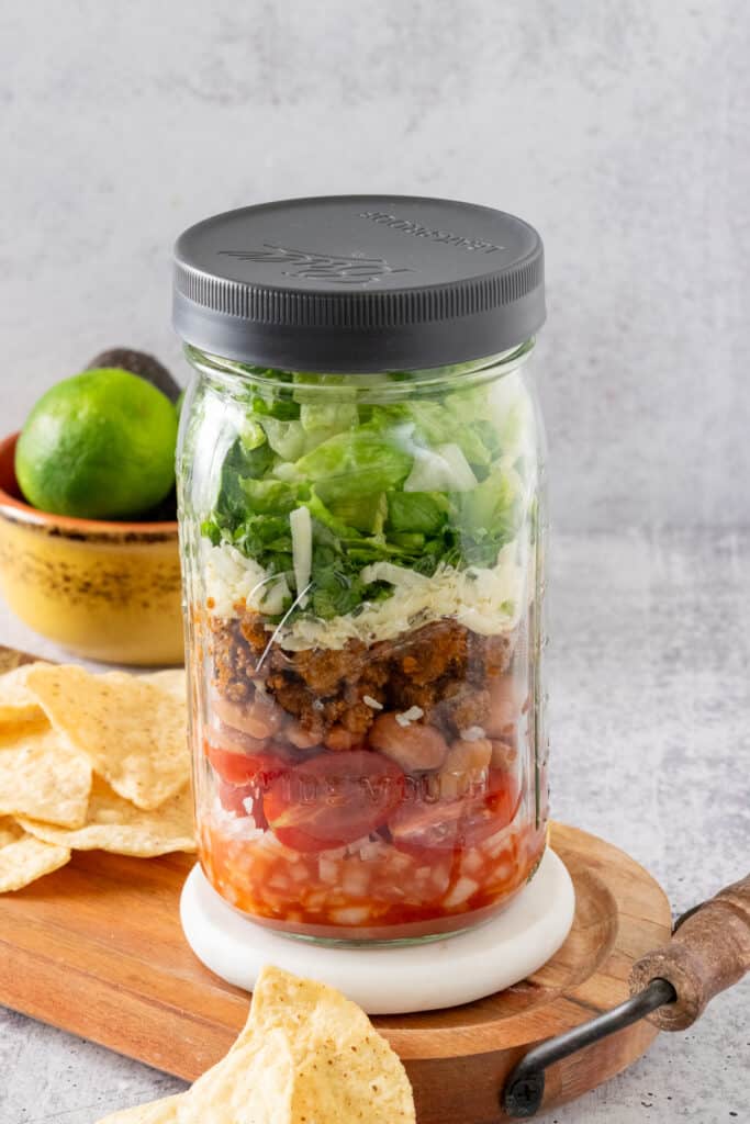 A layered mason jar taco salad surrounded by tortilla chips and an avocado and lime.