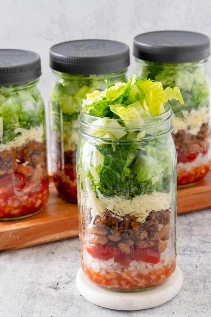 Four jars of taco salad in quart size mason jars, three with lids on top and one without a lid showing the lettuce popping out.