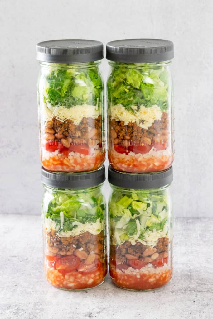 Four jars of taco salad stacked on top of each other in two rows showing all the layers of salad components packed in each jar.