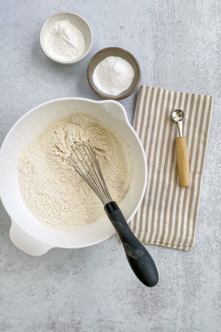 The dry buttermilk pancake mix ingredients being whisked together in a white bowl, which is next to small bowls of baking powder and baking soda.