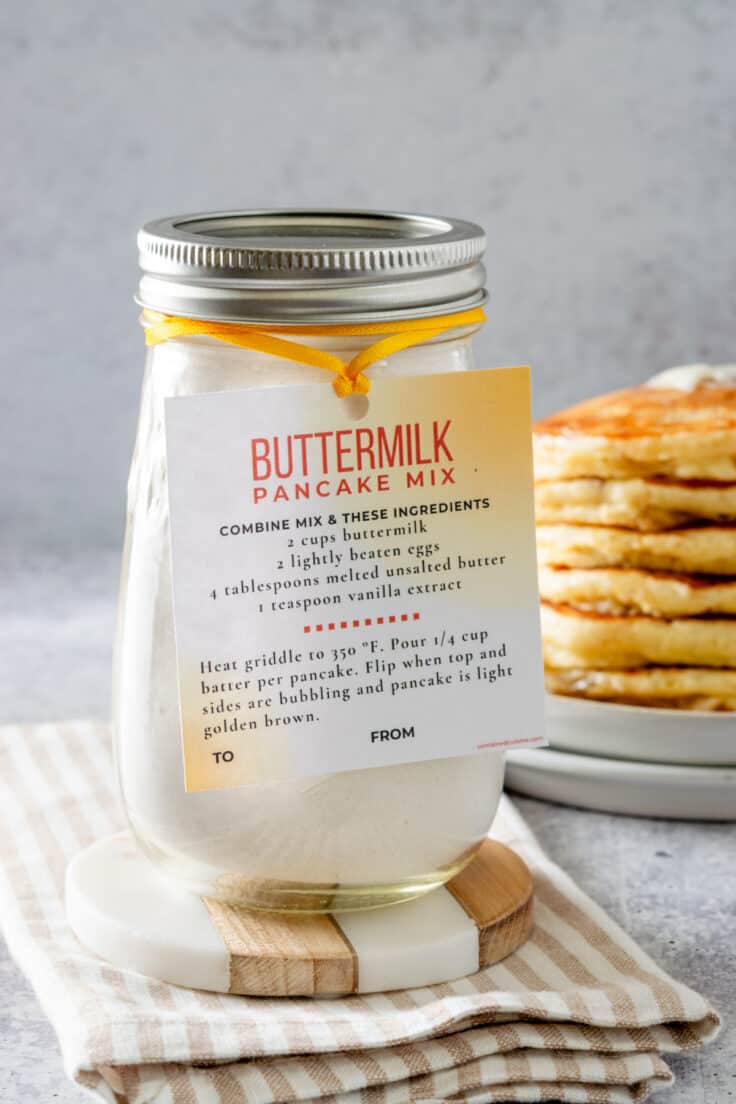Homemade buttermilk pancake mix in a mason jar with a gift tag tied on to it, sitting in front of a stack of buttermilk pancakes with butter on top and syrup dripping down the sides.
