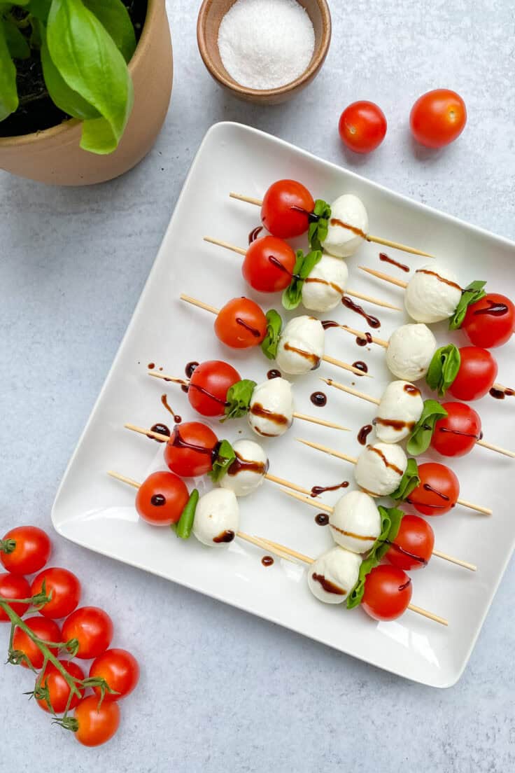 White serving platter with tomato, basil and mozzarella ball caprese skewers that have been drizzled with olive oil and balsamic glaze.