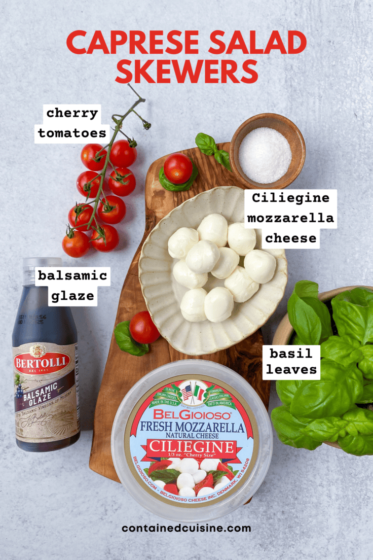 Overhead picture showing ingredients needed for making caprese appetizer skewers, including cherry tomatoes, ciliegine mini mozzarella cheese balls, basil leaves, olive oil, bottle of balsamic glaze and salt.