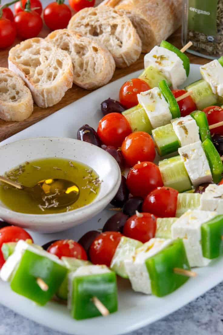 Greek salad skewer appetizers on a tray with a small bowl of olive oil and oregano, next to a board with crostini on it.