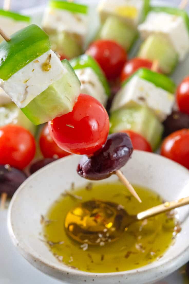 A Greek salad skewer held over a bowl of olive oil and oregano.