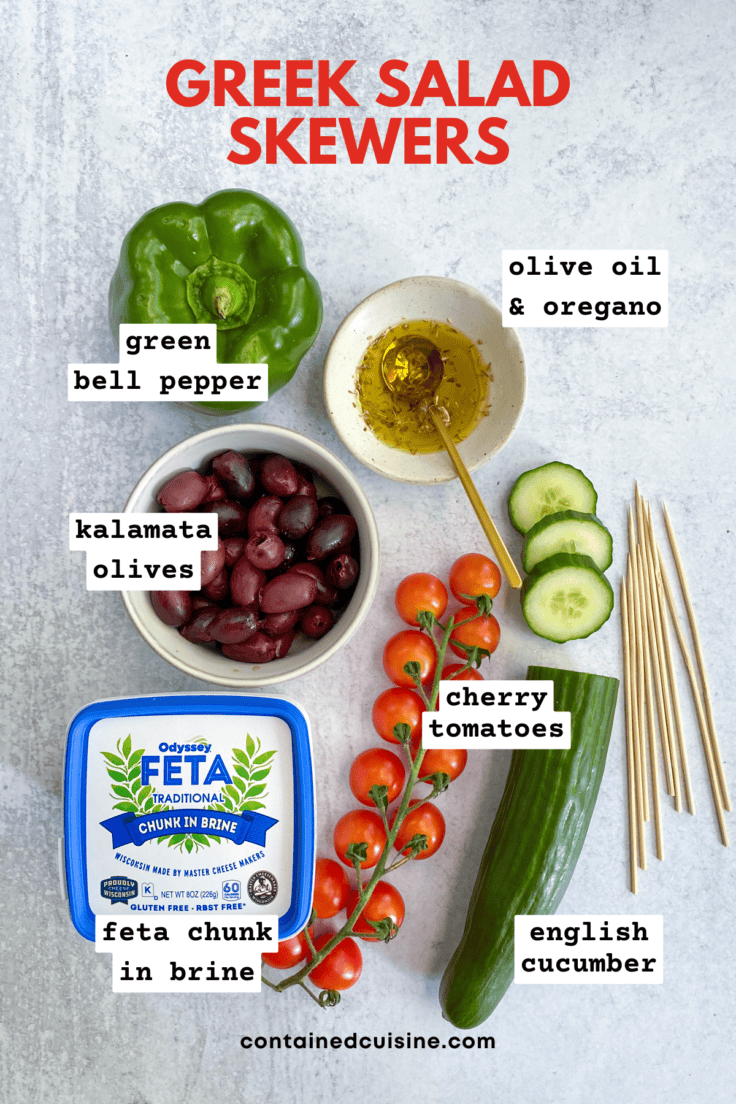 Overhead picture showing all the ingredients needed to make Greek salad skewers, including a green bell pepper, Kalamata olives. chunk feta cheese in brine, English cucumber, cherry tomatoes and bamboo sticks to skewer onto.