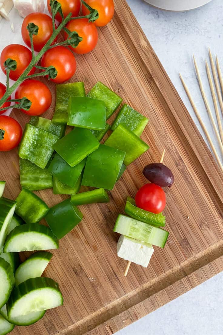 A cutting board with chopped ingredients and a single Greek salad skewer being assembled.