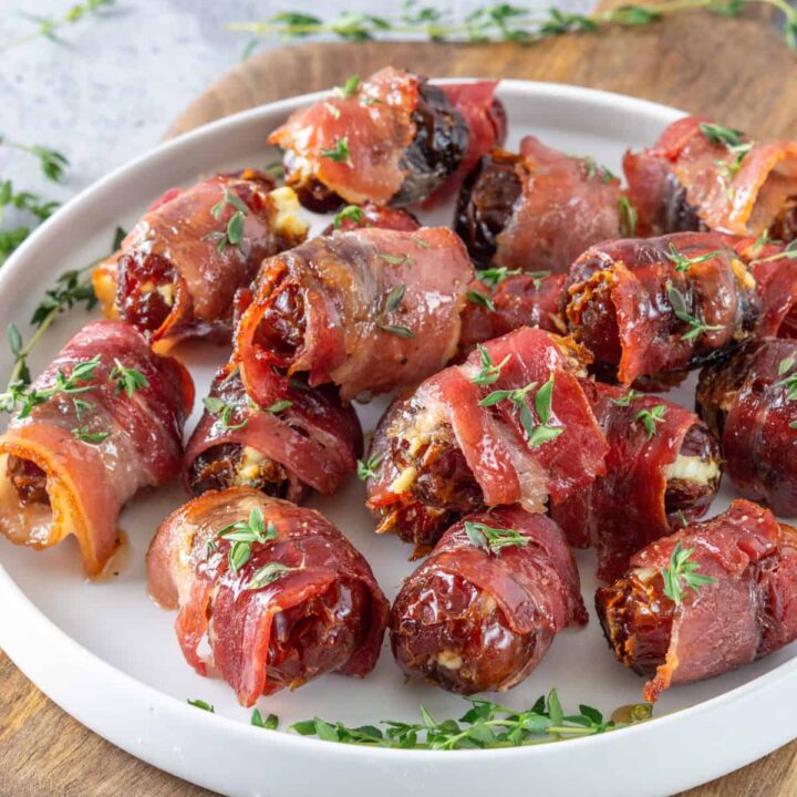 Pile of bacon-wrapped dates on a plate with maple glaze and fresh thyme sprinkled on top.