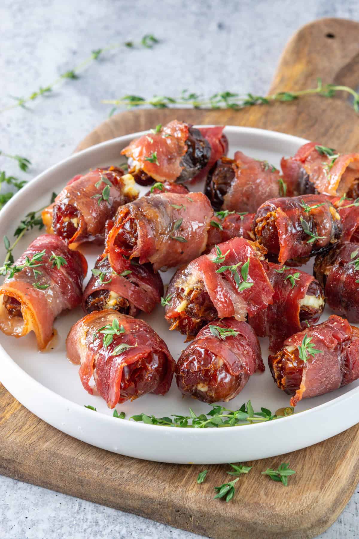 Pile of bacon-wrapped dates on a plate with maple glaze and fresh thyme sprinkled on top.