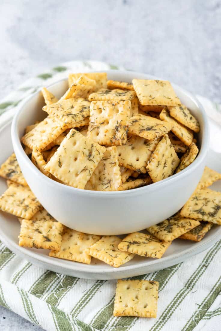 Bowl of mini saltine crackers that have been seasoned with dill pickle juice, dried dill, butter and spices, sitting on a green and white striped pot holder.