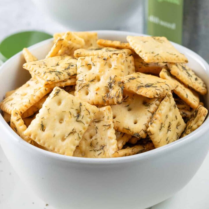 Bowl of homemade dill pickle crackers in front of a jar of dill pickles and bottle of dried dill spice.