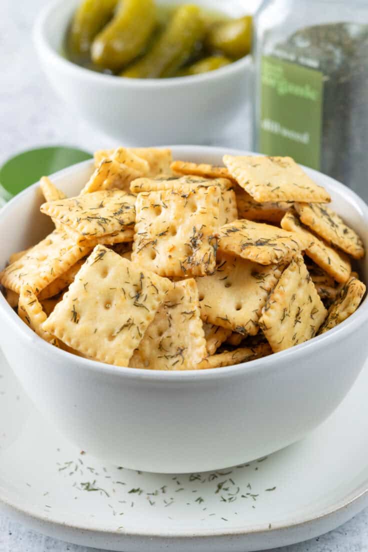 Bowl of homemade dill pickle crackers in front of a jar of dill pickles and bottle of dried dill spice.