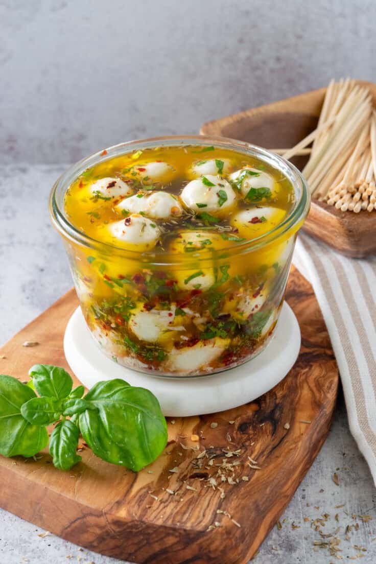 Ciliegine mozzarella balls in an olive oil and herb marinade. The cheese balls are in a small glass bowl next to some fresh basil leaves and a bowl of party picks for serving.