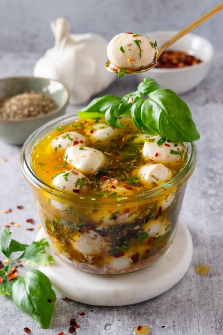 Glass bowl filled with marinated mozzarella balls in an olive oil and herb marinade with a small spoon lifting one of the pieces of cheese out.