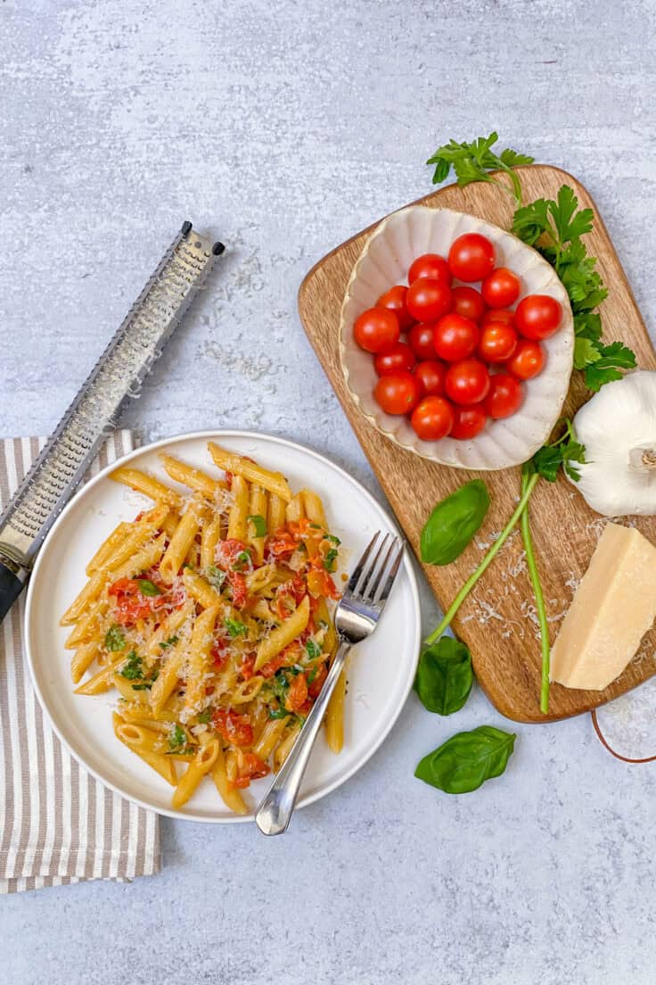 A dinner plate full of pasta with cherry tomato sauce and freshly grated parmesan on top. The plate has a fork on it and is next to a bowl full of cherry tomatoes, block of parmesan cheese and a few basil leaves.