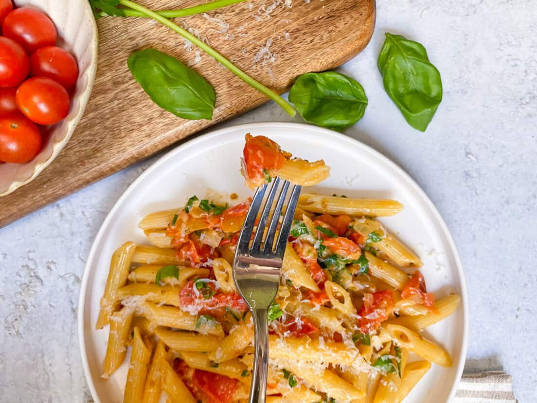A fork holding up penne pasta with cherry tomato sauce on it over a plate of pasta.
