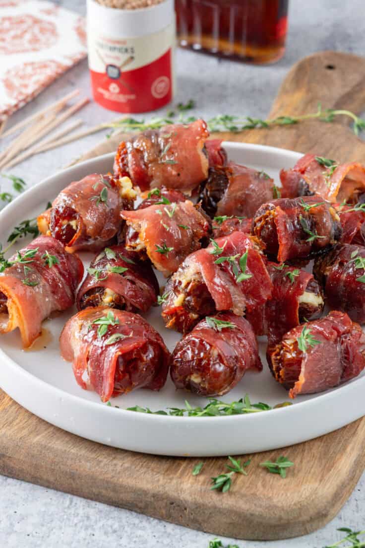 Bacon wrapped dates on a plate which is on a wooden board garnished with sprigs of thyme, next to a container of toothpicks and bottle of maple syrup.