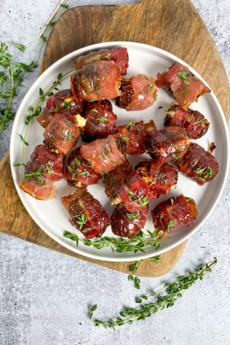 Overhead picture of a plate full of bacon wrapped dates appetizer which is sitting on a wood serving board with sprigs of thyme strewn about.