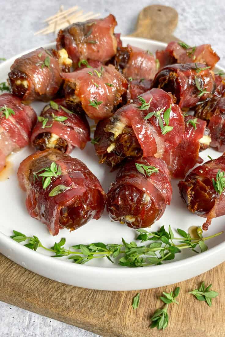 Close up view showing bacon wrapped dates glazed with maple syrup with fresh thyme sprinkled on top, on a plate which is on a wood board.