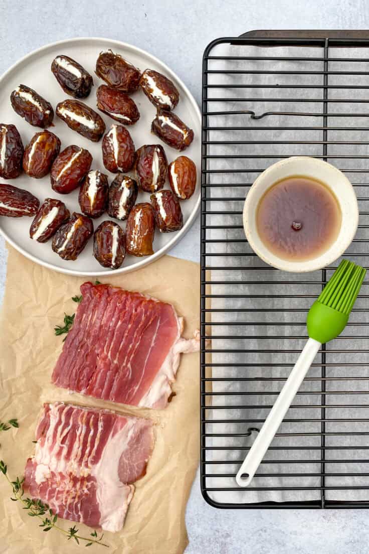 Strips of bacon cut in half lengthwise next to a plate full of goat cheese stuffed dates, as well as a bowl of maple syrup and pastry brush sitting on a rack and tray that's lined with parchment paper.