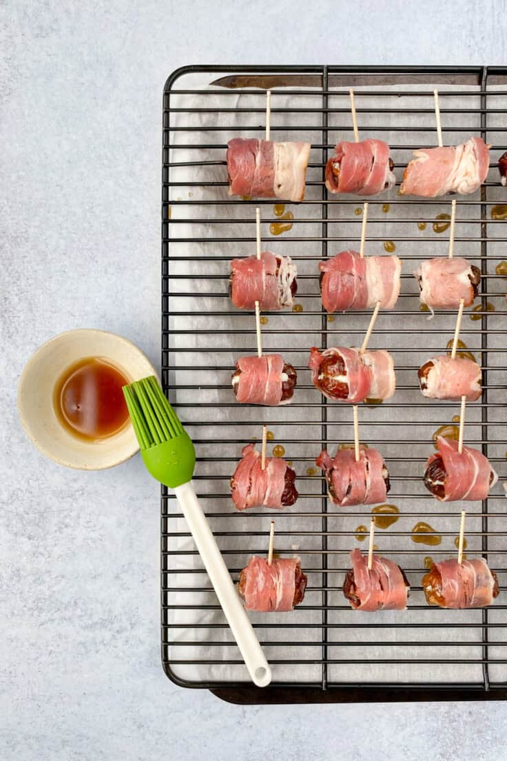 Maple syrup being brushed on to each bacon-wrapped date which have toothpicks inserted in each.