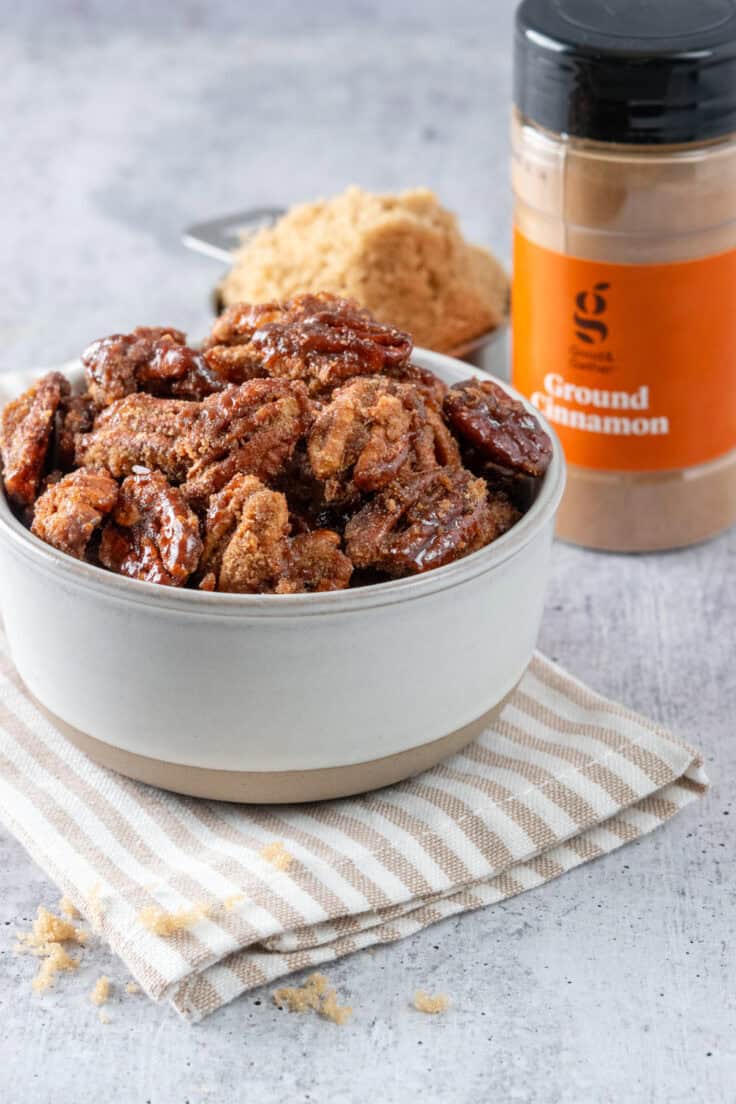 Bowl of homemade cinnamon sugar candied pecans next to a a bottle of ground cinnamon and a measuring cup overflowing with light brown sugar.