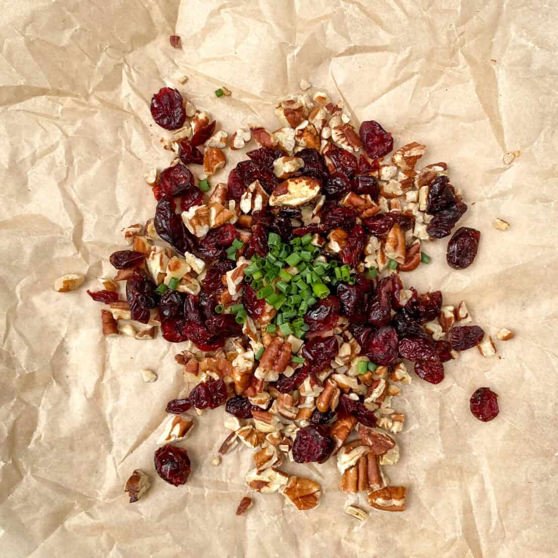 Piles of chopped pecans, dried cranberries and chives on a crinkled piece of brown parchment paper.