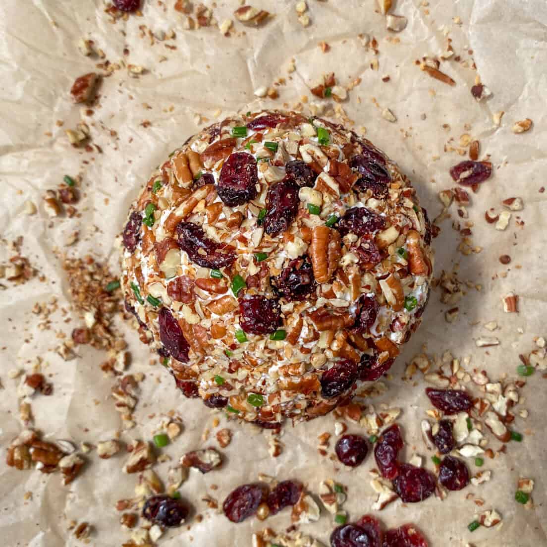 The cheese ball completely coated in the pecan and cranberry mixture with a little bit of extra nuts and berries on a piece of parchment paper around it.