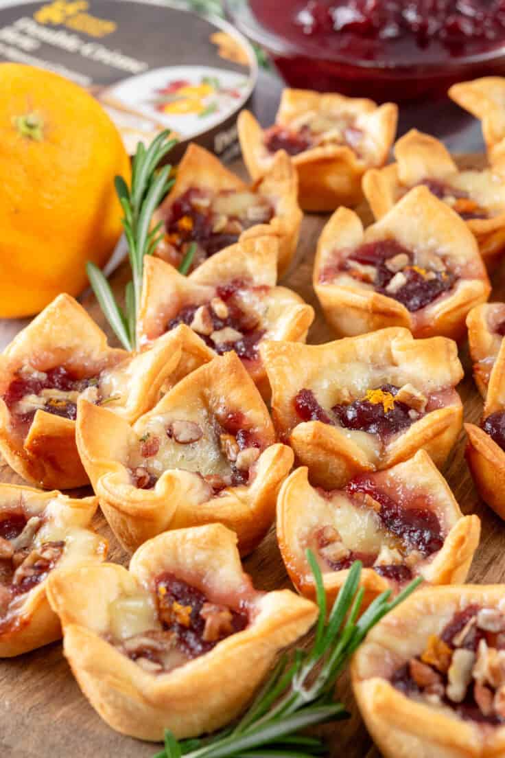 Cranberry brie bites on a board to serve as an appetizer next to a wheel of brie cheese, bowl of cranberry sauce and a zested orange.