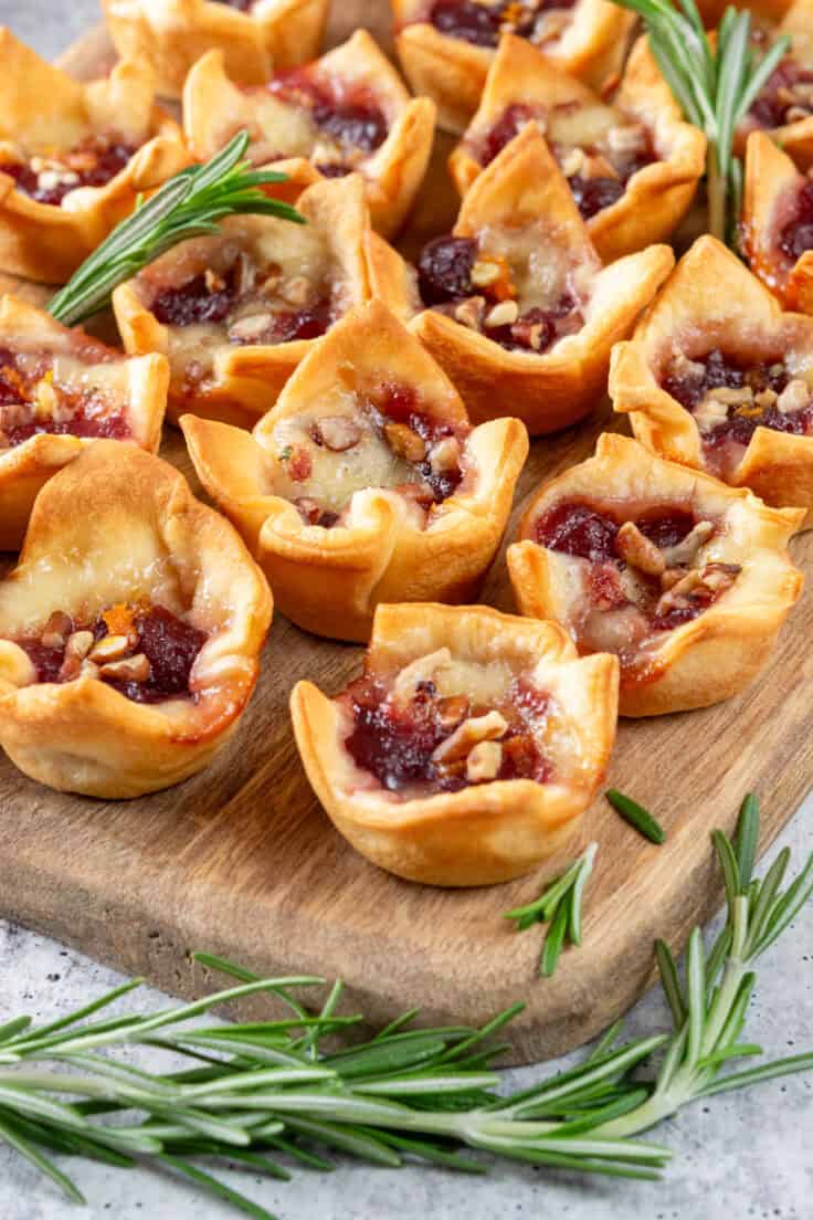 Cranberry brie bites on a board with sprigs of rosemary to garnish, showing the melted cheese and gooey cranberry sauce topped with orange zest and pecan chips.