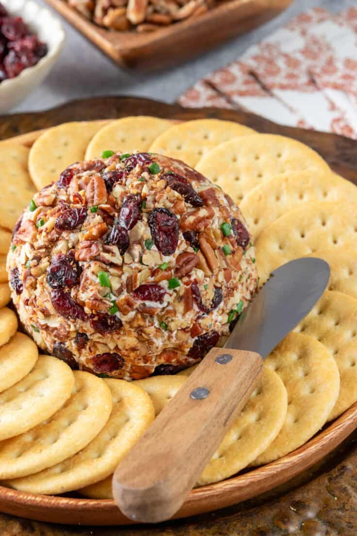 Cranberry pecan cheese ball set out as an appetizer on a board surrounded by crackers and a small knife for spreading the cheese onto crackers.