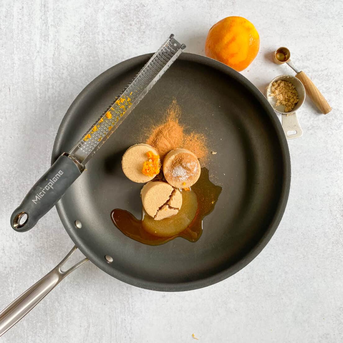 A skillet with all the ingredients in it, except the pecans, before they are heated and melted. There's an orange that's been zested and measuring spoon and cup next to the skillet.