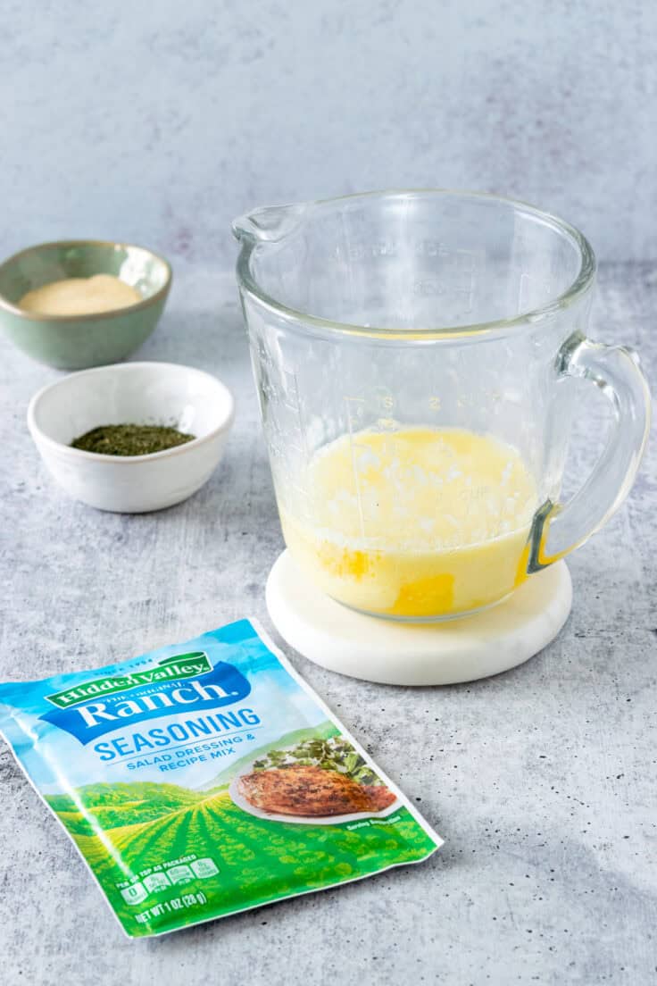 Melted butter in a heat-proof glass measuring cup, next to a packet of Hidden Valley ranch seasoning and bowls of dried dill and garlic powder.