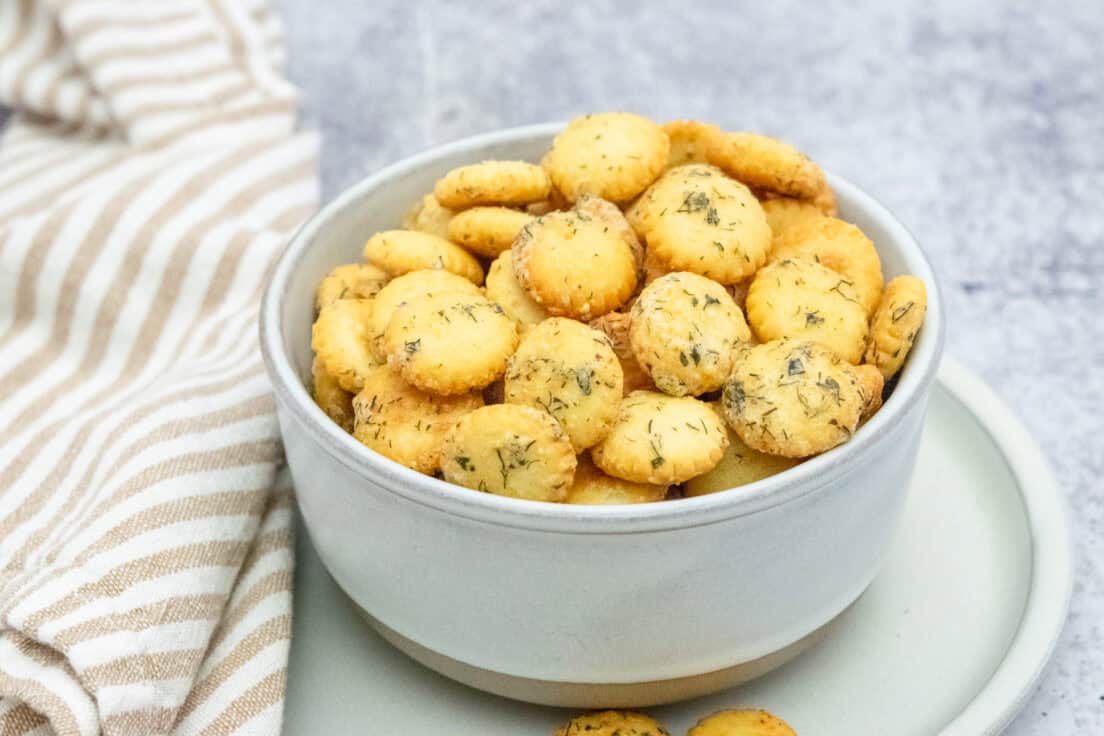 Ranch oyster crackers in a bowl which is sitting on a small plate, showing the dried dill and ranch seasoned baked onto the crispy crackers.