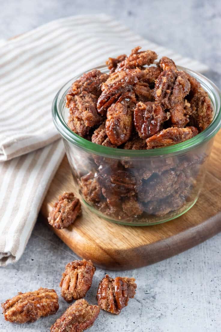 Glass jar full of homemade cinnamon sugar pecans. The jar is on a wooden board with more candied pecans on it.