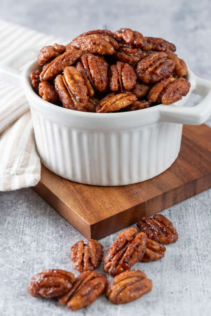 Small white bowl full of maple candied pecans, that is sitting on a wooden board with a few of the nuts scattered around.