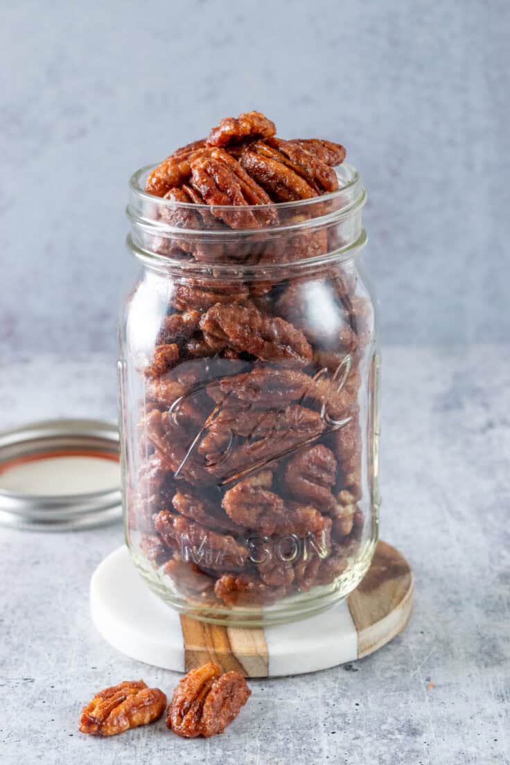 Maple candied pecans piled high in an open glass mason jar with the lid next to it.