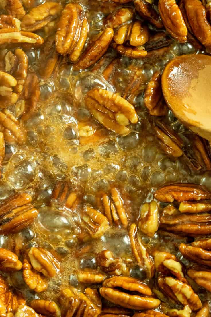 Candied pecans being stirred in a pan with a wooden spoon in a bubbling mixture of butter and maple syrup.